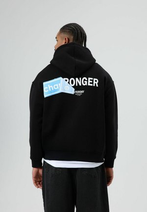 Zwarte hoodie met een grote witte tekst "STRONGER" op de achterkant, voorzien van een blauw rechthoekig label met de tekst "chay." Zachte stof, ontspannen pasvorm.