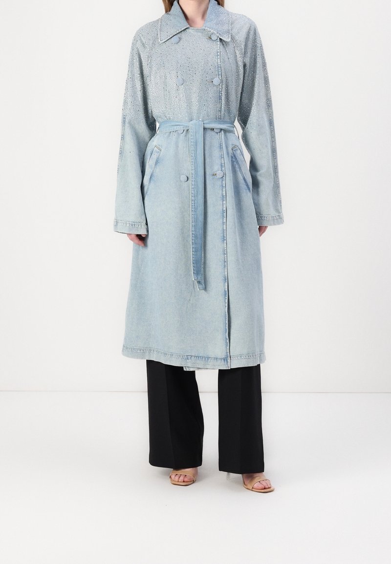 Femme portant un trench-coat en denim bleu clair ceinturé sur un pantalon noir à jambes larges et des talons beiges ouverts à l’avant, debout devant un fond blanc.