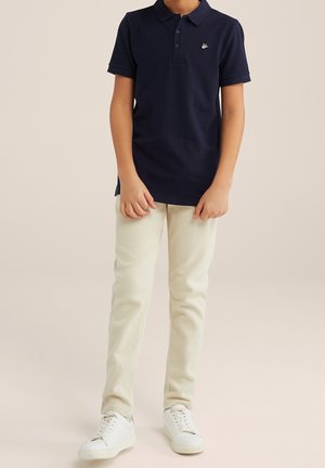 Polo blu navy a maniche corte, con colletto a tre bottoni e logo ricamato. Abbinato a pantaloni color crema e sneaker bianche.