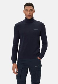 Maglione dolcevita blu navy con motivo a trama e maniche aderenti. Presenta un piccolo logo sul petto e un orlo a coste.