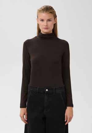 Femme aux cheveux blonds raides, vêtue d'un col roulé noir à manches longues et d'un pantalon noir taille haute, debout devant un fond uni.