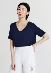 Blusa azzurro navy a maniche corte con scollo a V realizzata in tessuto morbido, abbinata a pantaloni bianchi con dettagli in pizzo intagliato sui lati.