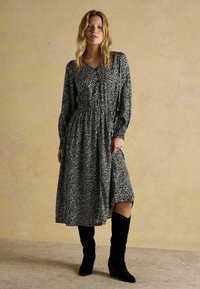 Frau in einem schwarz-weißen gemusterten Midikleid mit langen Ärmeln, kombiniert mit schwarzen kniehohen Stiefeln mit Absatz, steht auf einem strukturierten Boden.