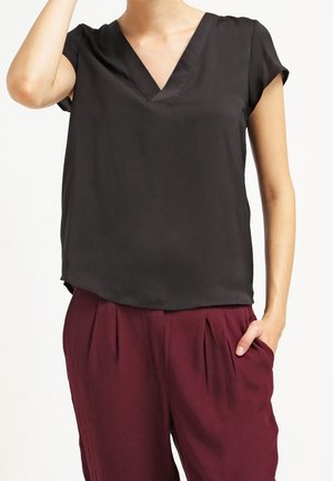 Woman wearing a black short-sleeve V-neck blouse and loose-fitting maroon pants with one hand in pocket.