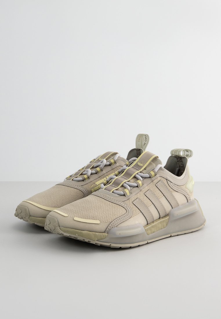 Zapatillas deportivas beige con una parte superior de malla texturizada, suela de goma y cordones grises. Presenta tres rayas características y una lengüeta trasera.