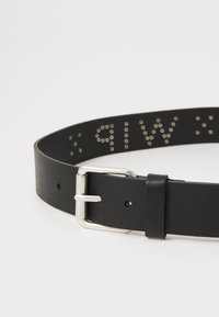 Sort læderbælte med en sølvmetal spænde. Har rækker af små metalnitter med bogstaver, der skaber et tekstureret design.