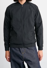 Svart idrottsjacka med dragkedja fram, ribbad mudd och midja. Har subtil märkning och sidofickor med en slät, matt textur.