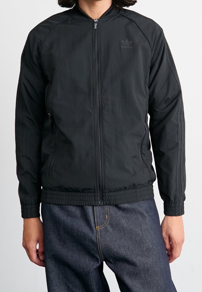 Svart idrottsjacka med dragkedja fram, ribbad mudd och midja. Har subtil märkning och sidofickor med en slät, matt textur.