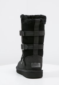 Botte en daim noir avec doublure en fourrure et sangles ajustables, ornée d'une plaque logo argentée UGG sur le côté ; design haut avec une finition texturée.