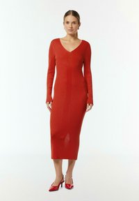 Rotes geripptes Strickkleid mit langen Ärmeln und V-Ausschnitt, figurbetonte Silhouette, knielang, kombiniert mit roten spitzen Absatzschuhen.