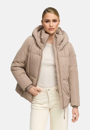 Vrouw in een taupe gewatteerde jas met capuchon over een lichtbeige ribbeltop en crèmekleurige broek met hoge taille, met één hand in haar zak.