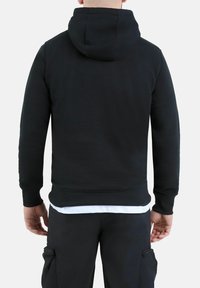 Persona che indossa un hoodie nero con cappuccio abbassato e pantaloni cargo neri, in piedi con la schiena rivolta verso lo spettatore su uno sfondo bianco.