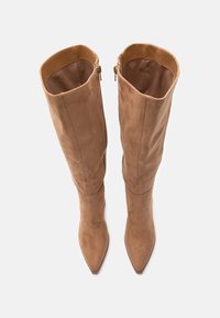 Una coppia di stivali alti fino al ginocchio in suede beige, con punte appuntite e cerniere laterali, visti dall'alto su uno sfondo bianco.