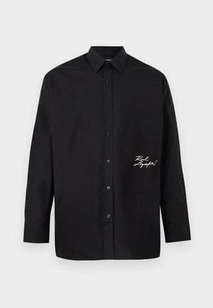 Chemise noire à manches longues avec col pointu et détail de signature cursive blanche sur le bas du côté droit avant.