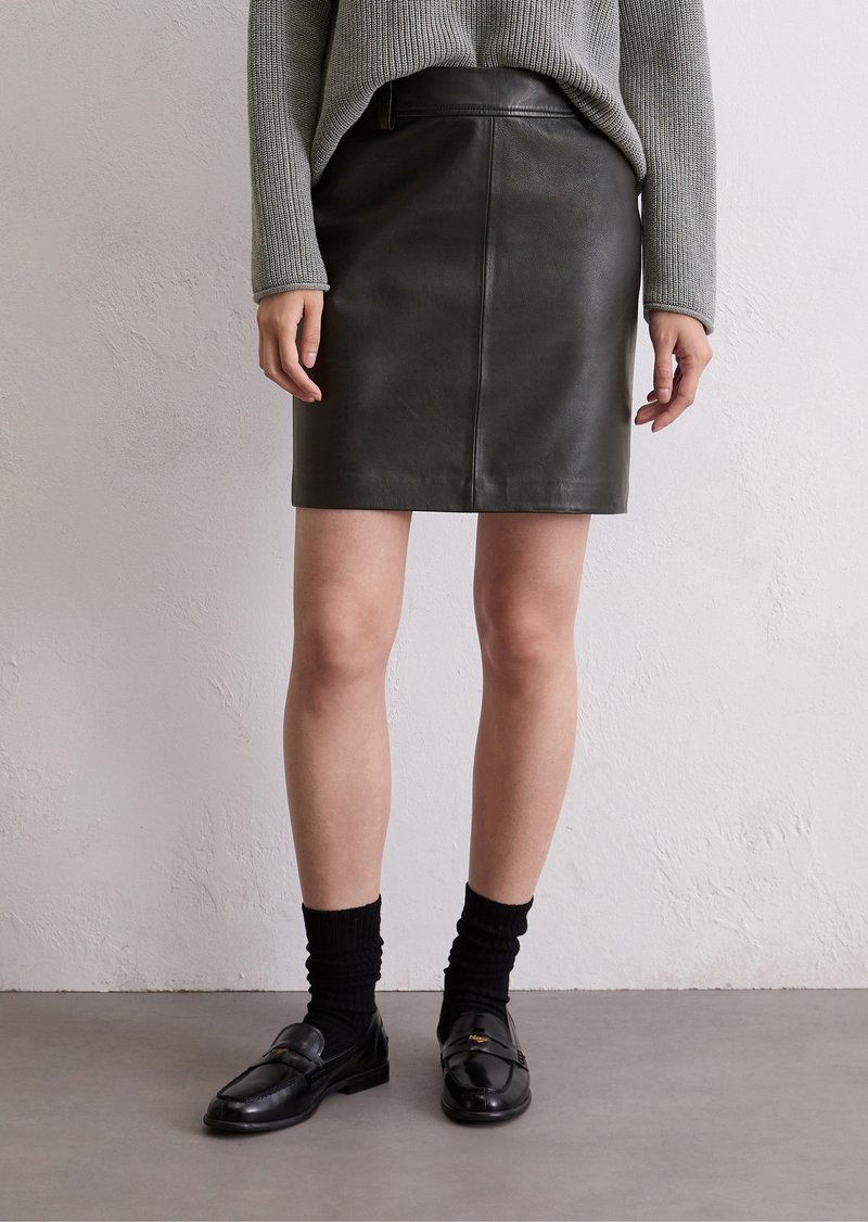 Zwarte leren rok met een rechte snit, voorzien van een voornaad detail. Gepaarde met een grijze gebreide trui en zwarte loafers met sokken.