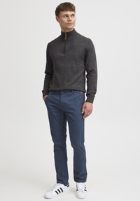 Un pull à col zippé gris foncé en tricot, associé à un pantalon bleu et à des baskets blanches avec des rayures noires. Design simple et moderne.