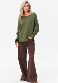 Oliwkowy sweter dzianinowy z szerokimi rękawami i ściągaczem, w połączeniu z brązowymi spodniami dzwonami. Całość charakteryzuje się luźnym krojem.