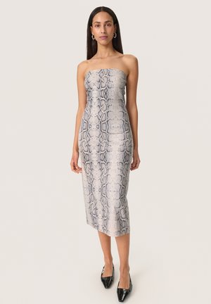 Mujer con vestido midi ajustado y sin tirantes con estampado de serpiente en gris y beige, combinado con tacones negros de punta afilada y pendientes de aro.