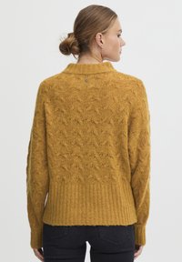 Senfgelbes Strickpullover mit strukturiertem Muster, geripptem Saum und Kragen. Rückseite hat ein kleines Logodetail nahe dem Hals.