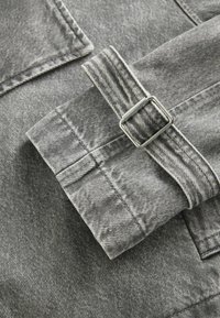 Manche de veste en jean grise avec sangle ajustable et détail de boucle rectangulaire argentée sur tissu texturé.