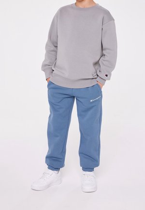 Felpa grigia, pantaloni della tuta blu, entrambi con il logo Champion. Le sneakers sono bianche. Il tessuto appare morbido con una vestibilità comoda e polsini a costine.