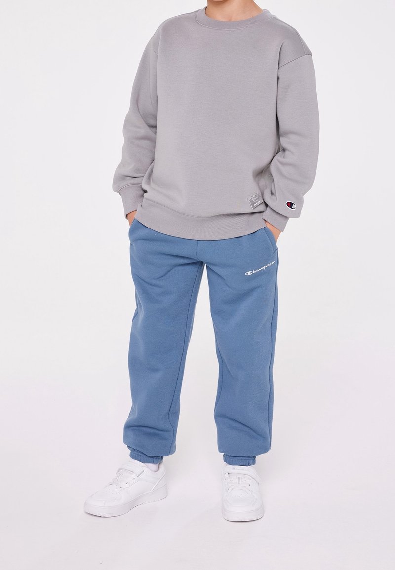 Felpa grigia, pantaloni della tuta blu, entrambi con il logo Champion. Le sneakers sono bianche. Il tessuto appare morbido con una vestibilità comoda e polsini a costine.