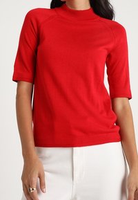 Červený pletený top s vysokým výstřihem, krátkým rukávem a přiléhavým střihem. Hladká textura, žádné viditelné vzory nebo ozdoby.