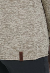 Pull en tricot de couleur crème et marron clair, avec un motif texturé. Comprend une étiquette en cuir marron au bas du côté gauche.
