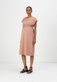 Maternity dress in soft blush fabric, featuring short cap sleeves, a gathered waistband, and a knee-length skirt. Paired with black strappy sandals.