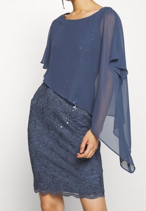 Kobieta ubrana w granatową sukienkę z koronkową spódnicą zdobioną cekinami i przezroczystymi, asymetrycznymi, warstwowymi rękawami, stojąca na jednolitym tle.