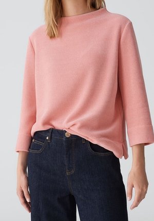 Personne portant un haut à manches longues côtelé rose clair et un jean bleu foncé, avec le haut légèrement rentré à l'avant.