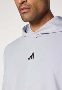 Felpa leggera grigia con tasca a marsupio, con un logo geometrico nero sul petto. Tessuto morbido, design casual e cappuccio regolabile con coulisse.