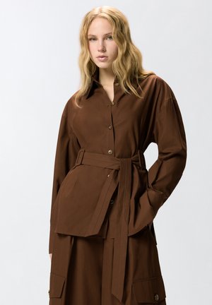 Jeune femme aux cheveux blonds ondulés portant une veste marron ceinturée avec boutons et un pantalon assorti avec poches, sur un fond uni.