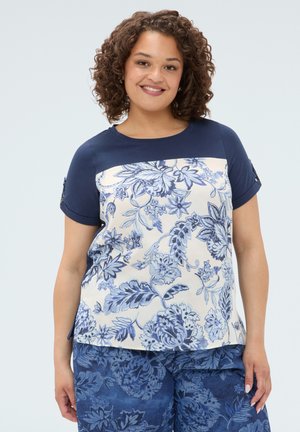 Mujer sonriente con el pelo rizado que lleva una blusa azul marino y blanca con estampado floral y pantalones azules con flores, de pie frente a un fondo liso.