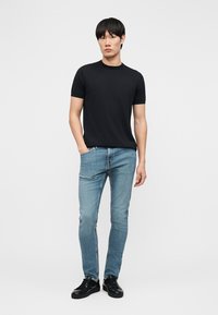 Młody mężczyzna stoi z rękami w kieszeniach niebieskich dżinsów, ubrany w jednolity czarny t-shirt z krótkim rękawem i czarne trampki na białym tle.