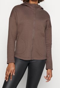 Sudadera marrón con cremallera hecha de tela texturizada, que incluye capucha, bolsillos laterales y bordes acanalados, combinada con mallas negras ajustadas.