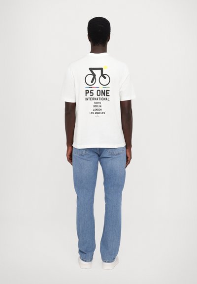 Camiseta blanca de algodón con un gráfico negro de una bicicleta y texto. Presenta líneas horizontales de colores. Combinada con jeans azul claro.
