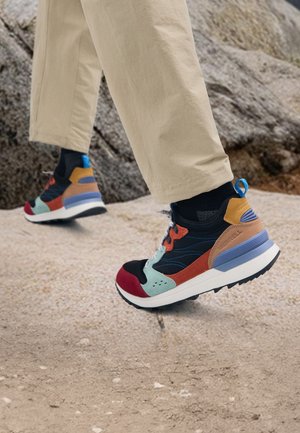 Person, die mehrfarbige Wanderschuhe und beige Hosen trägt und auf felsigem Gelände im Freien steht.