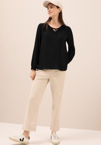 Blusa nera a maniche lunghe con apertura a feritoia e lacci al collo, abbinata a pantaloni beige chiaro a gamba larga e sneakers bianche.