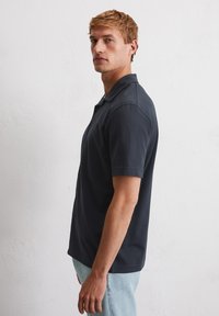 Man met kort blond haar, gekleed in een donkerblauw polo-shirt met korte mouwen en lichtblauwe jeans, staande in profiel tegen een witte muur.