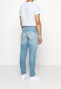 Světle modré džíny s tapered střih a přiznanou texturou, zkombinované se šedým tričkem a bílými teniskami. Zadní pohled ukazuje omezené detaily.