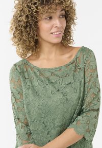 Top de encaje en verde apagado con mangas 3/4, que presenta patrones florales y un escote barco. Textura transparente y bordes de las mangas con forma de ondas.