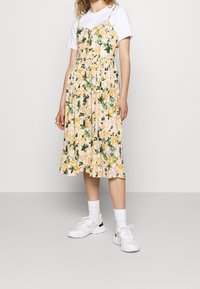 Vestido floral en amarillo y verde con un corsé ajustado, detalles de botones y una falda fluida; usado sobre una camiseta blanca y combinado con zapatillas blancas.