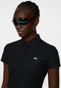 Czarna koszulka polo z teksturowanej tkaniny i zapięciem na guziki, z białym logo. Model nosi eleganckie czarne okulary przeciwsłoneczne.