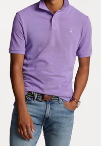 Man som bär en lavendelfärgad kortärmad polo med en liten logotyp, blå jeans, randigt bälte och brunt läderarmbandsur.