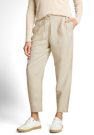 Woman wearing beige tapered pants with drawstring waist, light beige long-sleeve top, hands partially in pockets, and white sneakers with gum soles.