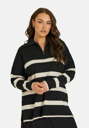 Kvinde med langt bølget hår iført en sort og hvid stribet langærmet sweater med en kort lynlås ved halsudskæringen.