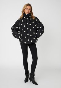 Polstret svart jakke med store hvite prikket, høy krage og frontlommer. Kombinert med svarte leggings og slouchy svarte støvler.