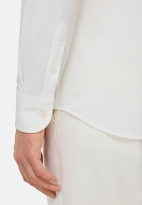 Bílá košile s dlouhými rukávy a manžetami na knoflíky, která se vyznačuje texturovanou tkaninou a přiléhavým střihem, a obsahuje jemně vyšívané logo blízko lemu.