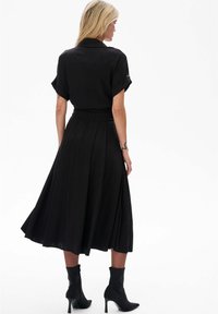 Zwart midi-jurk met korte opgerolde mouwen, geplooide rok, aansluitende taille en achterzakje. Gedragen met zwarte enkellaarsjes. Gladde stoffen textuur.